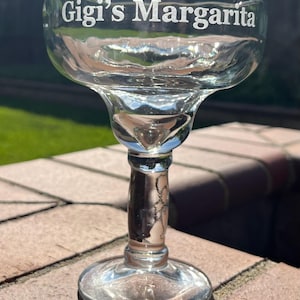 K&ouml;nnte beinhalten: Ein klares Glas Margarita-Glas mit einem breiten Rand und einem Stiel. Das Glas ist mit den Worten "Gigi's Margarita" graviert.