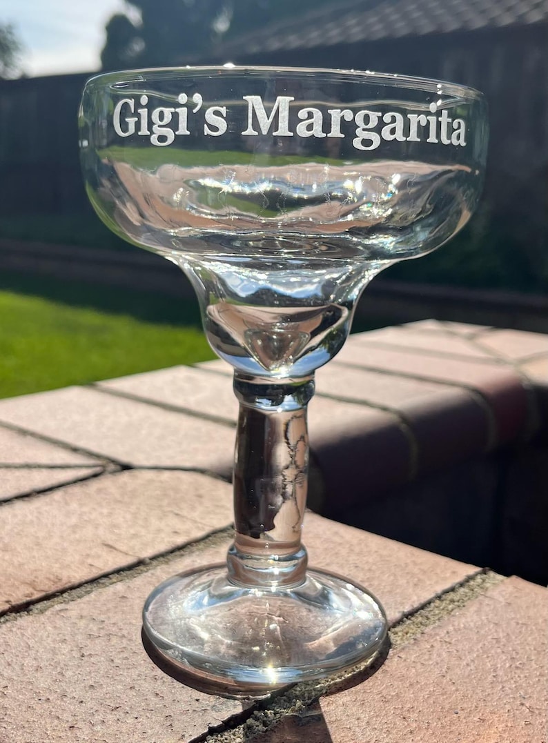 K&ouml;nnte beinhalten: Ein klares Glas Margarita-Glas mit einem breiten Rand und einem Stiel. Das Glas ist mit den Worten "Gigi's Margarita" graviert.