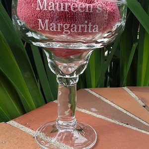 May include: A clear glass margarita glass with the words "Maureen's Margarita" etched on the side. The glass is sitting on a brick surface.