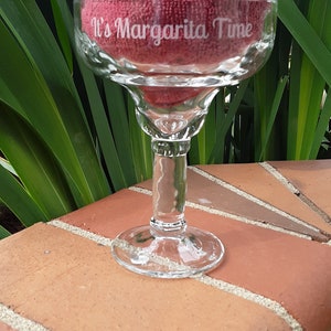 K&ouml;nnte beinhalten: Ein klares Glas Margarita-Glas mit dem Text "It's Margarita Time" auf der Seite. Das Glas steht auf einer Ziegelstein-Oberfl&auml;che.