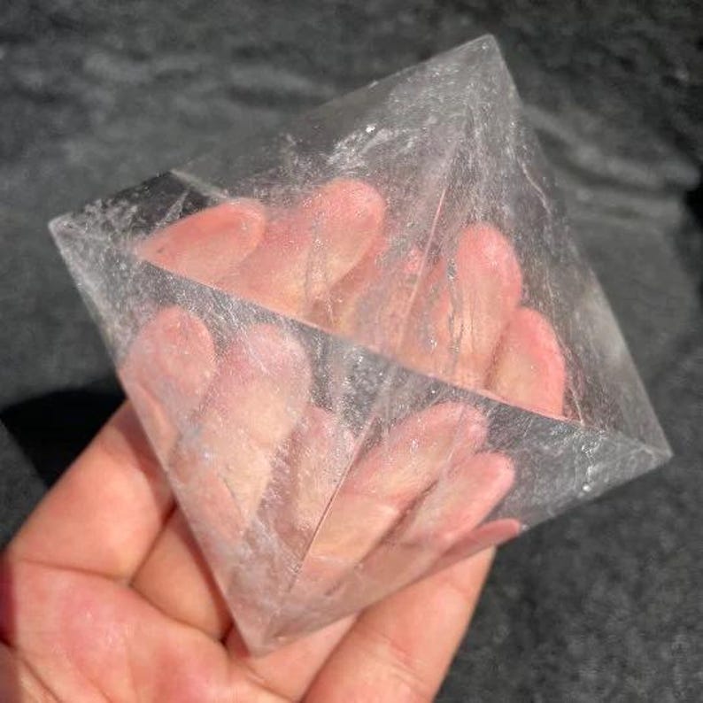 Puede incluir: Una pir&aacute;mide de cristal geom&eacute;trica transparente sostenida en una mano. La pir&aacute;mide es transparente, permitiendo ver la mano a trav&eacute;s del cristal. Los bordes de la pir&aacute;mide son afilados y bien definidos.
