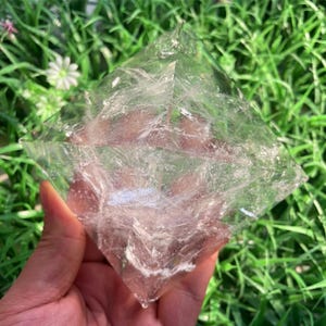 Puede incluir: Un cristal transparente en forma de pir&aacute;mide con vetas blancas internas. El cristal se sostiene en una mano sobre un fondo de hierba verde y peque&ntilde;as flores blancas. Las facetas del cristal capturan la luz, creando un efecto brillante.