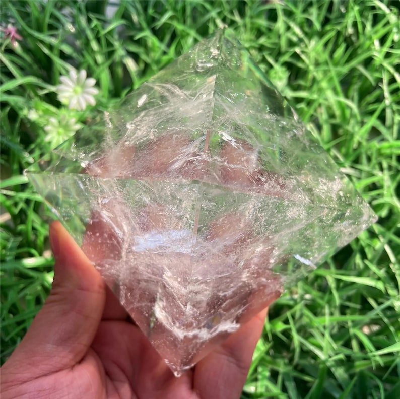 Puede incluir: Un cristal transparente en forma de pir&aacute;mide con vetas blancas internas. El cristal se sostiene en una mano sobre un fondo de hierba verde y peque&ntilde;as flores blancas. Las facetas del cristal capturan la luz, creando un efecto brillante.