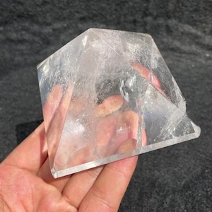 Puede incluir: Una pir&aacute;mide de cristal de cuarzo transparente con inclusiones internas visibles. La pir&aacute;mide se sostiene en una mano, mostrando su naturaleza transparente y forma geom&eacute;trica. El cristal parece ser un esp&eacute;cimen natural sin pulir.