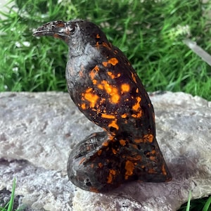 Könnte beinhalten: Eine handgefertigte Raben-Skulptur mit dunkler, strukturierter Oberfläche und leuchtend orangefarbenen Akzenten. Der Vogel sitzt auf einem Stein und zeigt seine detaillierten Merkmale. Dieses dekorative Stück ist ideal für die Inneneinrichtung.