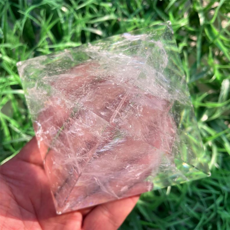 Puede incluir: Un cristal transparente en forma de pir&aacute;mide con un sutil tono rosado. El cristal se sostiene en una mano sobre un fondo de follaje verde. Las facetas de la pir&aacute;mide capturan la luz, creando un efecto brillante.