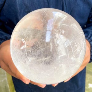 Peut inclure: Une sphère de cristal de quartz clair, tenue dans les mains, reflétant la lumière. La sphère est un orbe parfait, présentant des inclusions naturelles et des motifs internes. Cette boule de cristal décorative est un objet de décoration unique.