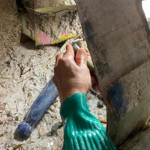 Puede incluir: Una persona con un guante verde utiliza una herramienta para trabajar en una piedra. La piedra est&aacute; rodeada de escombros. Un objeto azul est&aacute; en el fondo.