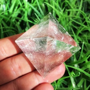 Puede incluir: Un cristal transparente en forma de pir&aacute;mide con un tinte rosado. El cristal se sostiene en una mano sobre un fondo de hierba verde. La pir&aacute;mide tiene una forma geom&eacute;trica con facetas visibles e inclusiones internas.