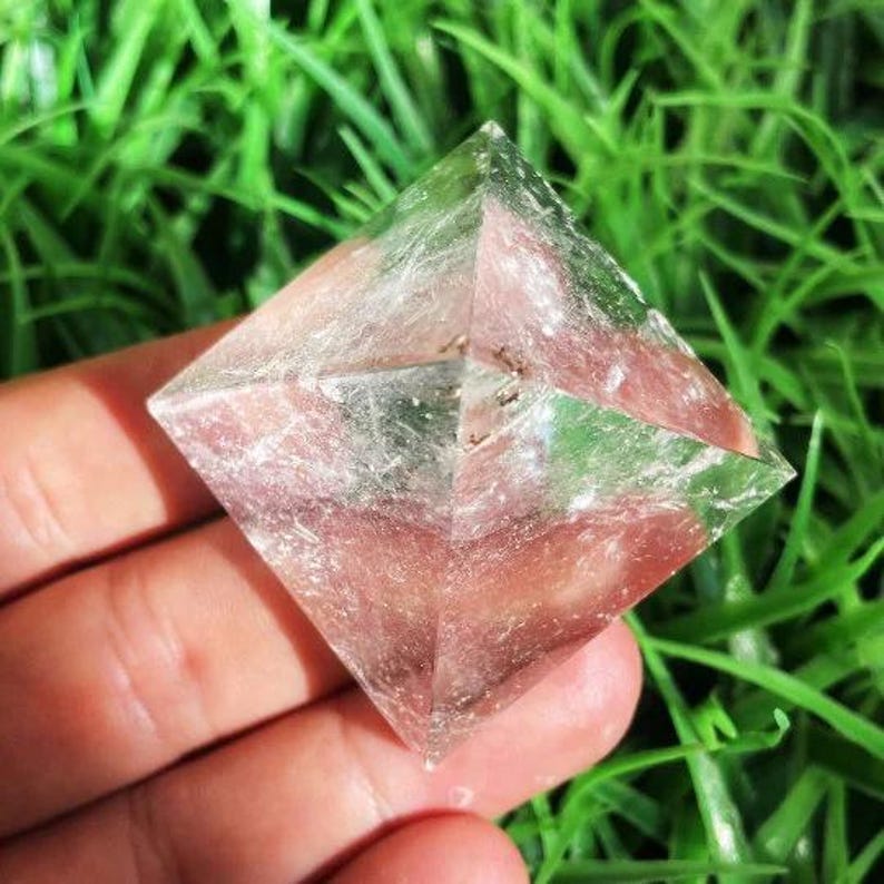 Puede incluir: Un cristal transparente en forma de pir&aacute;mide con un tinte rosado. El cristal se sostiene en una mano sobre un fondo de hierba verde. La pir&aacute;mide tiene una forma geom&eacute;trica con facetas visibles e inclusiones internas.