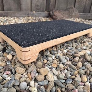 May include: A wooden step stool with a black, speckled top. The stool has a rectangular shape with rounded corners and four short legs. The wood has a natural finish, and the step stool is designed for use in various settings.