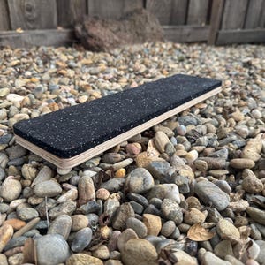 May include: A rectangular balance board with a black, speckled top surface and a light-coloured wooden frame. The board is designed for exercise and is approximately 76 cm long.
