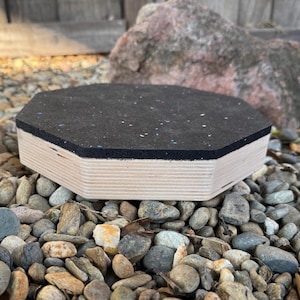 May include: A hexagonal wooden platform with a black rubber top, resting on a bed of small stones. The platform's sides are made of light-colored wood, and the rubber top has flecks of color. The background includes a large rock and a wooden fence.