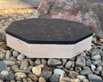 May include: A hexagonal wooden platform with a black rubber top, resting on a bed of small stones. The platform's sides are made of light-colored wood, and the rubber top has flecks of color. The background includes a large rock and a wooden fence.