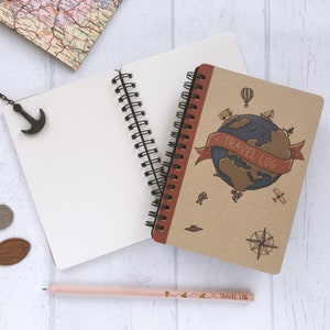 Può includere: Un diario di viaggio marrone con una rilegatura a spirale e un'illustrazione di un globo terrestre. Il diario ha il testo "TRAVEL LOG" sulla copertina. Il diario è aperto su una pagina bianca e una matita è appoggiata accanto.