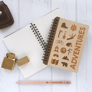 Può includere: Un quaderno marrone con una rilegatura a spirale bianca e le parole "ADVENTURES" stampate sulla copertina. Il quaderno è aperto per rivelare pagine vuote. Nell'immagine sono presenti altri oggetti, tra cui un accendino, una matita e una piccola borsa marrone.