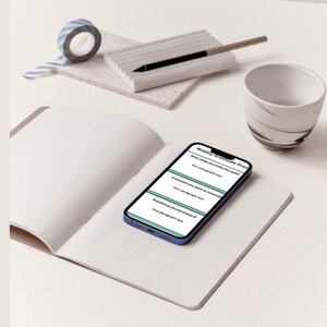 K&ouml;nnte beinhalten: Ein offenes Notizbuch mit leeren Seiten, ein Smartphone mit einer "Weekly gratitude Prompt", ein Stift, eine kleine Keramiktasse und eine Rolle blau-wei&szlig; gestreiftes Washi-Tape. Die Gegenst&auml;nde sind auf einer hellen Oberfl&auml;che angeordnet.
