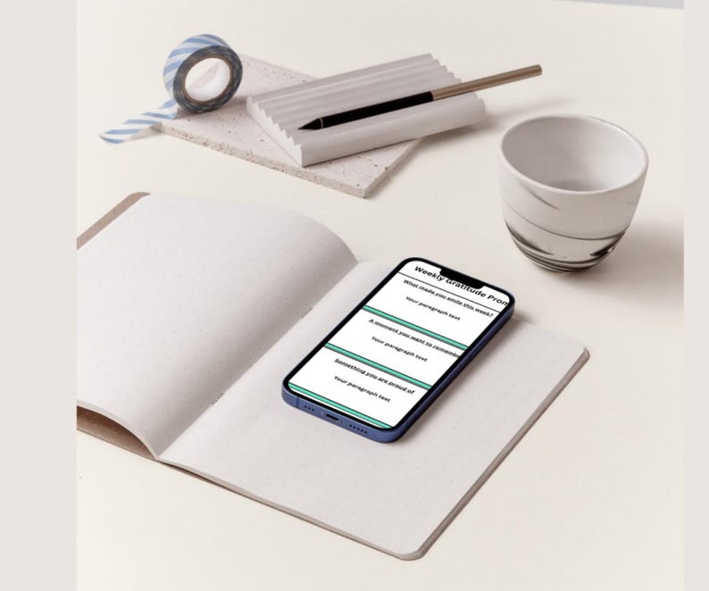 K&ouml;nnte beinhalten: Ein offenes Notizbuch mit leeren Seiten, ein Smartphone mit einer "Weekly gratitude Prompt", ein Stift, eine kleine Keramiktasse und eine Rolle blau-wei&szlig; gestreiftes Washi-Tape. Die Gegenst&auml;nde sind auf einer hellen Oberfl&auml;che angeordnet.