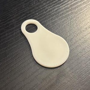 Può includere: Un piccolo tag a forma di goccia, bianco sporco, con un foro circolare nella parte superiore. Il tag è appoggiato su una superficie di legno scuro. Un oggetto rosso e circolare è visibile sullo sfondo.