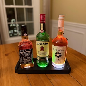 Könnte beinhalten: Drei beleuchtete Spirituosenflaschen auf einem schwarzen Tablett. Die Flaschen sind Jack Daniel's Tennessee Whiskey, Jameson Irish Whiskey und Skrewball Peanut Butter Whiskey. Die Flaschen werden von unten beleuchtet und erzeugen eine farbenfrohe Anzeige.