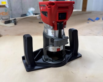 Puede incluir: Una fresadora Milwaukee roja y plateada con base y asas negras. La herramienta eléctrica está diseñada para trabajar la madera y se muestra sobre una superficie de madera de color claro.