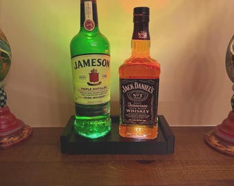 Puede incluir: Dos botellas de whisky, Jameson y Jack Daniel's, sobre una bandeja negra. La botella de Jameson es verde con tapón rojo, y la de Jack Daniel's ámbar con etiqueta negra. Sobre una superficie de madera.