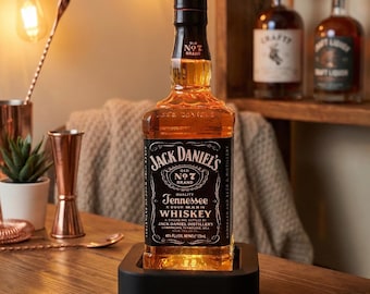 Puede incluir: Una botella de Jack Daniel's Tennessee Whiskey, exhibida sobre una superficie de madera. La botella está en un soporte negro, con la etiqueta visible. La imagen está iluminada con luces de cadena cálidas, creando un ambiente acogedor.