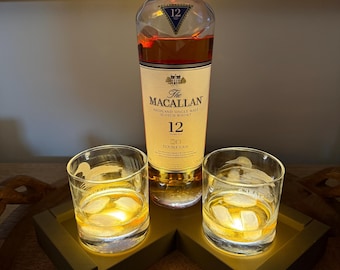 Puede incluir: Una botella de whisky escocés The Macallan 12 Double Cask con dos vasos de whisky y hielo en una bandeja de madera. La botella tiene una etiqueta dorada y los vasos están iluminados.