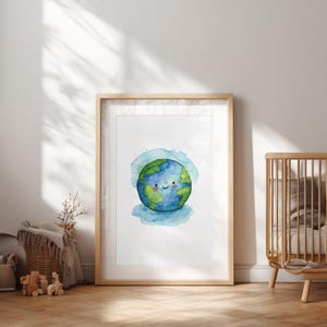 Pode incluir: Uma impressão em aquarela emoldurada de uma Terra sorridente com oceanos azuis e continentes verdes. A obra de arte está em uma moldura de madeira clara e é exibida em um ambiente de berçário, com um berço visível ao fundo.