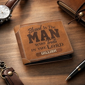 May include: A brown leather wallet with the text "Blessed Is The MAN Who Trusts In The Lord" and the name "William" engraved on the front. The wallet is surrounded by a watch, pen, keychain, and notebook on a wooden surface.