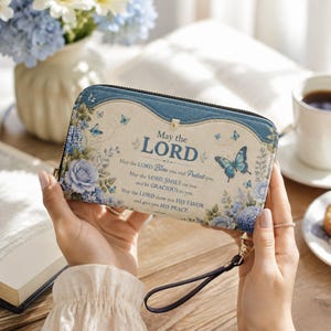 May include: A rectangular zip-around wallet with a cream-colored front featuring blue floral patterns, butterflies, and the text: May the LORD Bless you and Protect you. May the LORD SMILE on you and be GRACIOUS to you. May the LORD show you HIS FAVOR and give you HIS PEACE.