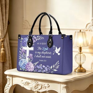 Op de afbeelding: Een paarse tote bag met zwarte handvatten, met een wit kruis, bloemaccenten en de tekst "HE IS RISEN The Lord is my shepherd; I shall not want. Psalm 23:1". De tas heeft een decoratief ontwerp met een duif en een muzieknoot.