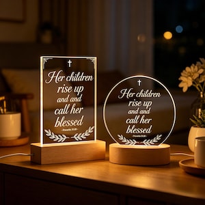 Op de afbeelding: Twee verlichte acryl nachtlampjes met houten voet. Eén is rechthoekig, de andere cirkelvormig. Beide tonen de tekst "Her children rise up and call her blessed" met een kruis en bloemmotief. De lampjes staan op een houten oppervlak.