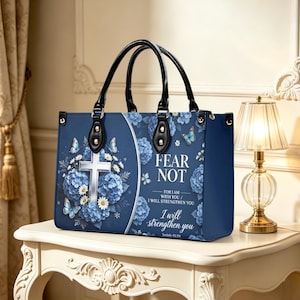 Puede incluir: Un bolso tote azul marino con asas negras. El bolso presenta una cruz blanca, detalles florales azules y mariposas. El texto en el bolso dice "FEAR NOT" y "I will strengthen you". El bolso está sobre una mesa blanca.