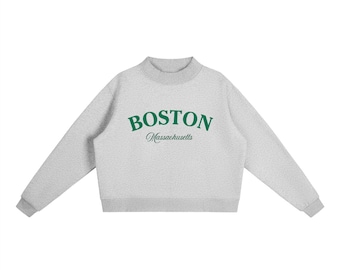 Op de afbeelding: Een lichtgrijs sweatshirt met het woord "BOSTON" in groene letters op de borst, met "Massachusetts" eronder gedrukt. De sweatshirt heeft een ronde hals en lange mouwen met geribde manchetten.