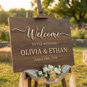Puede incluir: Un letrero de bienvenida de boda de madera sobre un caballete. El letrero dice "Bienvenida a la boda OLIVIA & ETHAN 24 de agosto de 2026". Un arreglo floral y tela cubren la parte inferior del letrero. El fondo incluye balas de heno y un jardín.