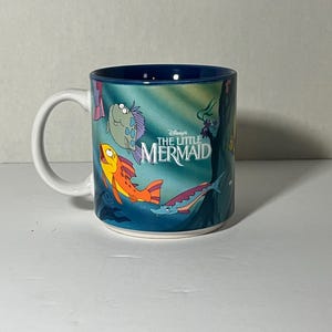 May include: A white ceramic mug featuring characters from Disney's The Little Mermaid. The mug has a blue interior and a wraparound design with cartoon fish and the movie's logo. The handle is white.