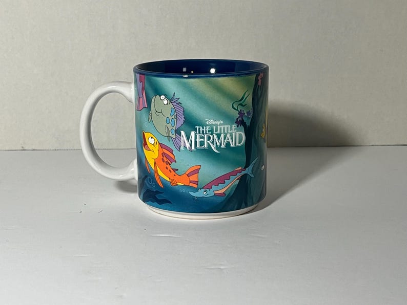 May include: A white ceramic mug featuring characters from Disney's The Little Mermaid. The mug has a blue interior and a wraparound design with cartoon fish and the movie's logo. The handle is white.