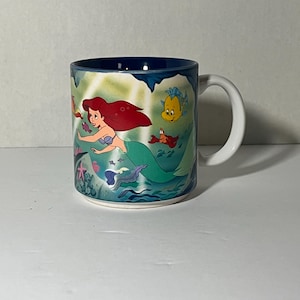 May include: A ceramic mug featuring a colorful illustration of Ariel from The Little Mermaid. The design includes Ariel with red hair, a blue seashell bra, and a green tail, swimming with Flounder and Sebastian. The mug has a white handle and a dark blue interior.