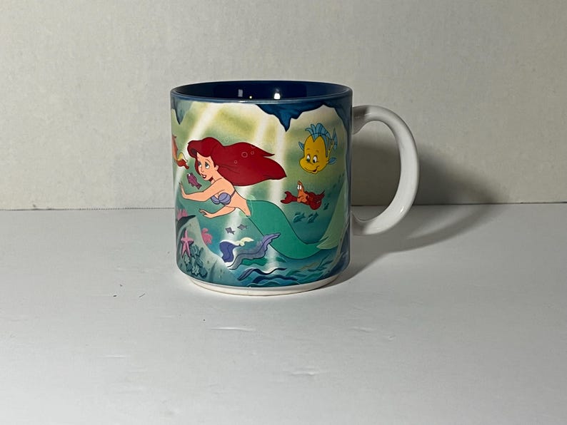 May include: A ceramic mug featuring a colorful illustration of Ariel from The Little Mermaid. The design includes Ariel with red hair, a blue seashell bra, and a green tail, swimming with Flounder and Sebastian. The mug has a white handle and a dark blue interior.