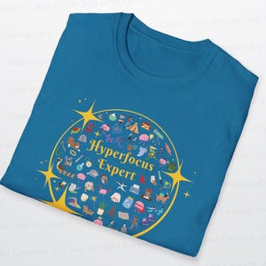 Może przedstawiać: Turkusowa koszulka z okrągłym, złotym wzorem. Wz&oacute;r zawiera słowa "Hyperfocus Expert" otoczone małymi, kolorowymi ilustracjami i gwiazdkami.