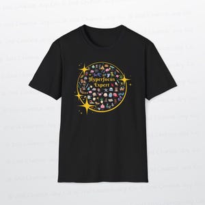 Może przedstawiać: Czarny t-shirt z okrągłym, złotym wzorem z napisem "Hyperfocus Expert". Projekt zawiera r&oacute;żne małe, kolorowe ilustracje i akcenty gwiazd. Codzienny, swobodny t-shirt.