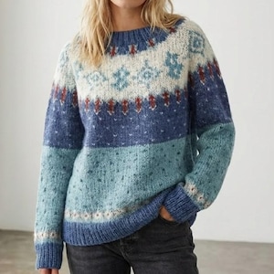 May include: A blue, white, and red patterned sweater. The sweater features a round neckline and long sleeves. The design includes a band of snowflakes and diamond shapes. The sweater is made of a soft, knitted material.