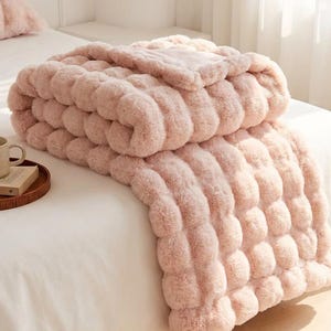 Puede incluir: Un juego de manta y almohada de piel sintética rosa claro. La manta está doblada sobre una cama blanca, con una taza y un libro en una bandeja de madera en primer plano. La manta tiene un diseño texturizado y acanalado.