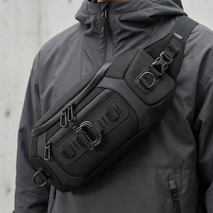 May include: Black crossbody bag with multiple zippered compartments and adjustable strap. The bag features a sleek, modern design with metal hardware and a durable, textured material. The bag is worn across the body.