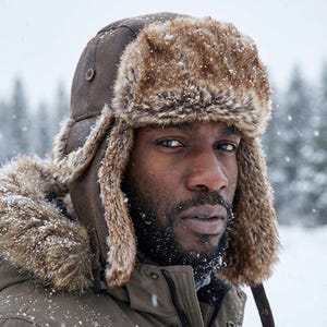 Op de afbeelding: Een bruine pilotenmuts met imitatiebont. De hoed heeft oorflappen en een knoopsluiting. De persoon op de foto draagt de hoed in een besneeuwde omgeving. De hoed maakt deel uit van een winterkledingcollectie.