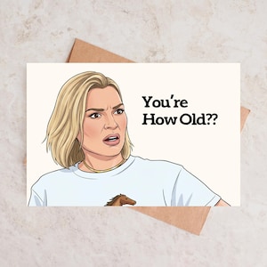 May include: Greeting card featuring a cartoon illustration of a woman with blonde hair and a surprised expression. The card has the text "You're How Old??". The woman is wearing a light blue t-shirt with a horse graphic.