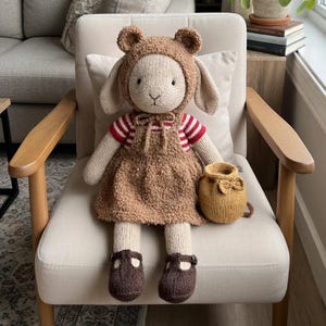 May include: A handmade, knitted doll with a bear-eared hood, brown dress, and matching shoes. The doll has long, floppy ears and is holding a small, knitted purse. It is sitting on a cream-colored chair with wooden arms.