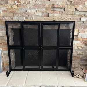 May include: A black metal fireplace screen with a mesh design. The screen has two doors and is set against a stone brick wall. A photo album and stuffed dog toys are on the hearth.