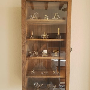 May include: A wooden display cabinet with glass doors, showcasing miniature glass and metal figurines. The cabinet is mounted on a wall and contains various detailed objects, including a bicycle, a typewriter, and a telephone.